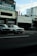 Several taxis are parked alongside a street in front of a commercial building with signage in Japanese. The sky is partially cloudy, and the overall setting gives an urban feel.