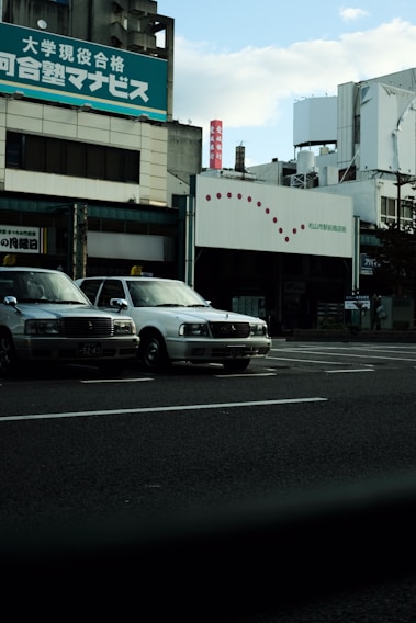 Several taxis are parked alongside a street in front of a commercial building with signage in Japanese. The sky is partially cloudy, and the overall setting gives an urban feel.