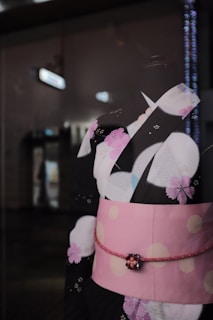 A traditional kimono with a black base adorned with pink and white cherry blossom patterns. It features a pink obi with white dots and a decorative tie at the waist. The background is dimly lit, suggesting an indoor setting.