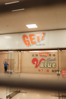 A closed shop or market is visible with a large sign reading 'GET!' above and another shuttered door marked with 'Value Fresh'. A person wearing a blue shirt is seen inside near the entrance, possibly cleaning or inspecting.