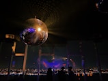 A large, reflective disco ball hangs from the ceiling, casting light patterns across the room. Below, silhouettes of people are visible against a backdrop of a panoramic nighttime cityscape. The scene is dimly lit, with colorful lights creating a festive and dynamic atmosphere.