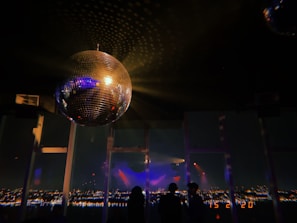 A large, reflective disco ball hangs from the ceiling, casting light patterns across the room. Below, silhouettes of people are visible against a backdrop of a panoramic nighttime cityscape. The scene is dimly lit, with colorful lights creating a festive and dynamic atmosphere.