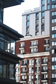 A view of urban architecture featuring several multi-story buildings with modern designs. The buildings have a mix of red brick and gray panels, with numerous windows reflecting light. Shadows play across the facades, and some balconies are visible.