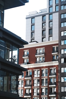 A view of urban architecture featuring several multi-story buildings with modern designs. The buildings have a mix of red brick and gray panels, with numerous windows reflecting light. Shadows play across the facades, and some balconies are visible.