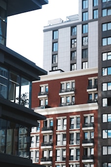 A view of urban architecture featuring several multi-story buildings with modern designs. The buildings have a mix of red brick and gray panels, with numerous windows reflecting light. Shadows play across the facades, and some balconies are visible.