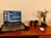 A laptop displaying a chat application is placed on a wooden desk. To the right, there are two small potted plants and a taller plant in a glass vase. A wireless mouse rests on the desk alongside a small dark bottle.