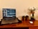 A laptop displaying a chat application is placed on a wooden desk. To the right, there are two small potted plants and a taller plant in a glass vase. A wireless mouse rests on the desk alongside a small dark bottle.