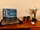 A laptop displaying a chat application is placed on a wooden desk. To the right, there are two small potted plants and a taller plant in a glass vase. A wireless mouse rests on the desk alongside a small dark bottle.