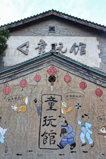 The image features a mural on a traditional-style building with Asian architecture. The mural includes cartoonish characters and red lanterns. There are children and fox illustrations interacting with each other, with some speech bubbles containing Chinese characters. The background of the mural is a textured wall with a light tan color, while the architecture above the mural has a classic tiled roof and gray brickwork.