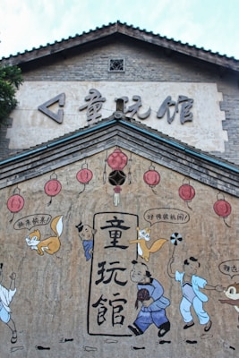 The image features a mural on a traditional-style building with Asian architecture. The mural includes cartoonish characters and red lanterns. There are children and fox illustrations interacting with each other, with some speech bubbles containing Chinese characters. The background of the mural is a textured wall with a light tan color, while the architecture above the mural has a classic tiled roof and gray brickwork.