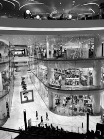 A multi-level shopping mall featuring modern architecture with sleek, curving lines. The image depicts an interior view with a prominent clothing store, mannequins dressed in outfits, and several shoppers walking along the marble floor. Overhead lights create a contemporary and elegant atmosphere.