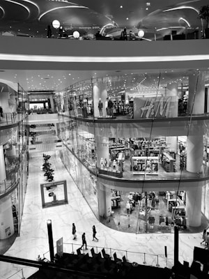A multi-level shopping mall featuring modern architecture with sleek, curving lines. The image depicts an interior view with a prominent clothing store, mannequins dressed in outfits, and several shoppers walking along the marble floor. Overhead lights create a contemporary and elegant atmosphere.