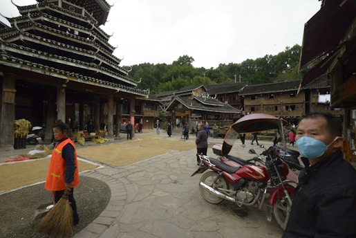 A traditional village scene with people walking and performing daily activities in an open area surrounded by wooden structures. A person is wearing an orange vest and holding a broom, while another individual nearby is wearing a blue face mask. A motorcycle with a canopy is parked in the foreground, and the architecture features wooden buildings with tiered roofs, typical of traditional styles. Trees can be seen in the background.