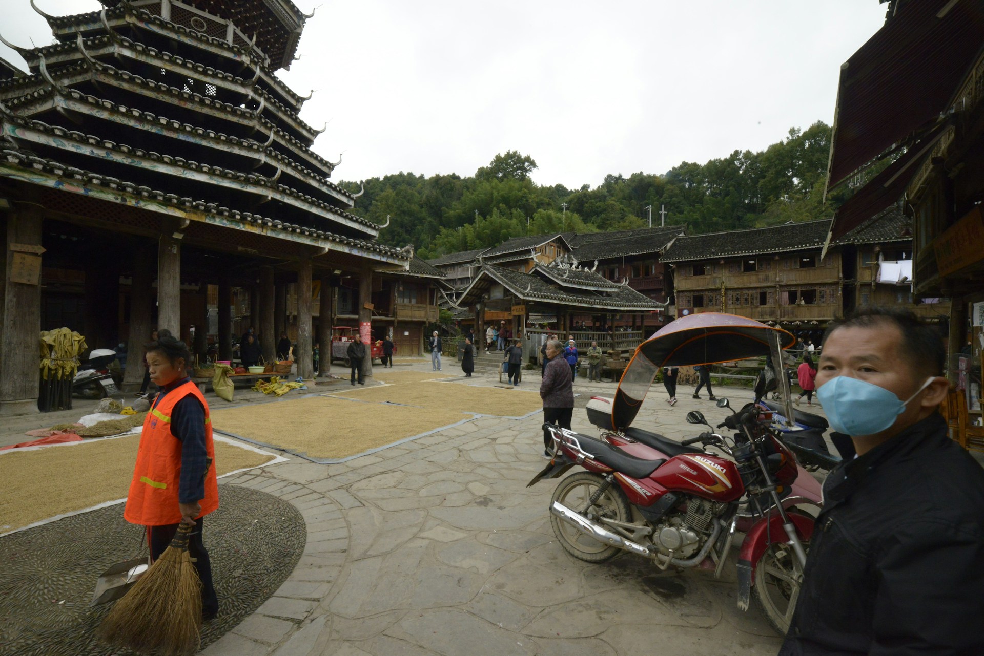 A traditional village scene with people walking and performing daily activities in an open area surrounded by wooden structures. A person is wearing an orange vest and holding a broom, while another individual nearby is wearing a blue face mask. A motorcycle with a canopy is parked in the foreground, and the architecture features wooden buildings with tiered roofs, typical of traditional styles. Trees can be seen in the background.