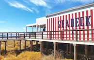 A seaside restaurant with bold red and white vertical stripes on its exterior. There is a wooden boardwalk leading to the entrance. The sky is clear and blue, and sparse vegetation grows below the boardwalk.