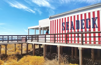 A seaside restaurant with bold red and white vertical stripes on its exterior. There is a wooden boardwalk leading to the entrance. The sky is clear and blue, and sparse vegetation grows below the boardwalk.