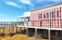A seaside restaurant with bold red and white vertical stripes on its exterior. There is a wooden boardwalk leading to the entrance. The sky is clear and blue, and sparse vegetation grows below the boardwalk.