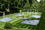 A beautifully manicured garden featuring multiple rectangular and circular water fountains, surrounded by neatly trimmed green lawns and dense trees. The water jets rise gracefully from the fountains, creating a serene and elegant atmosphere.
