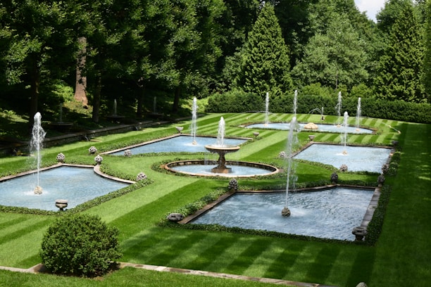 A beautifully manicured garden featuring multiple rectangular and circular water fountains, surrounded by neatly trimmed green lawns and dense trees. The water jets rise gracefully from the fountains, creating a serene and elegant atmosphere.