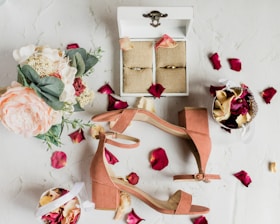 A flat lay of wedding accessories including a pair of peach-colored heeled sandals, a floral bouquet with pink and white flowers, and a white box with rings inside. Red and cream-colored rose petals are scattered around, with a small basket filled with similar petals.
