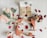 A flat lay of wedding accessories including a pair of peach-colored heeled sandals, a floral bouquet with pink and white flowers, and a white box with rings inside. Red and cream-colored rose petals are scattered around, with a small basket filled with similar petals.