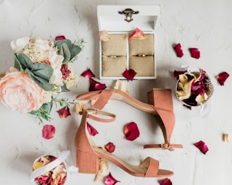 A flat lay of wedding accessories including a pair of peach-colored heeled sandals, a floral bouquet with pink and white flowers, and a white box with rings inside. Red and cream-colored rose petals are scattered around, with a small basket filled with similar petals.