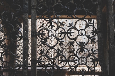 Intricate wrought iron patterns form ornate designs with floral and circular motifs. The metalwork features a rustic, aged appearance, with light filtering through behind, highlighting the craftsmanship.
