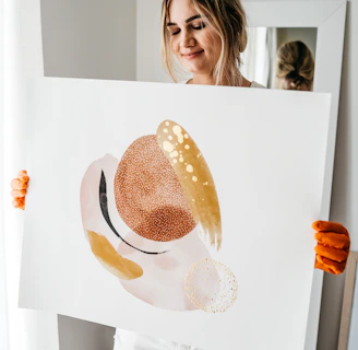 A person with a gentle expression holds up an abstract painting. The painting features organic shapes in hues of brown, beige, and gold, with a texture that resembles brush strokes and dots. The person wears casual white shorts and orange gloves, standing in a softly lit room with neutral decor.