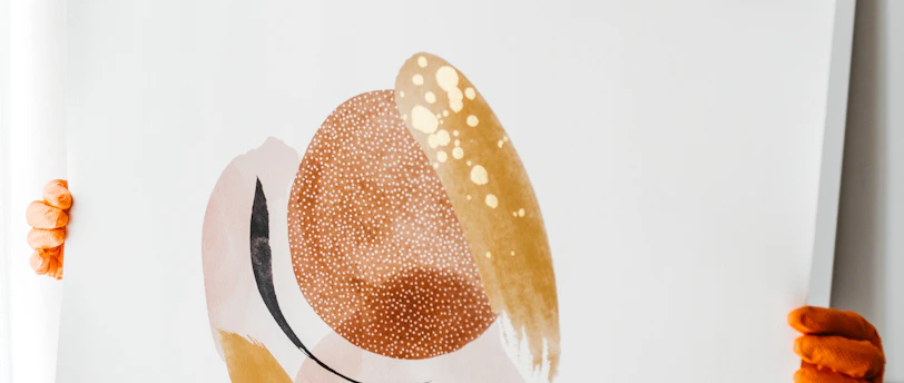A person with a gentle expression holds up an abstract painting. The painting features organic shapes in hues of brown, beige, and gold, with a texture that resembles brush strokes and dots. The person wears casual white shorts and orange gloves, standing in a softly lit room with neutral decor.