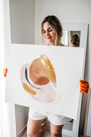 A person with a gentle expression holds up an abstract painting. The painting features organic shapes in hues of brown, beige, and gold, with a texture that resembles brush strokes and dots. The person wears casual white shorts and orange gloves, standing in a softly lit room with neutral decor.