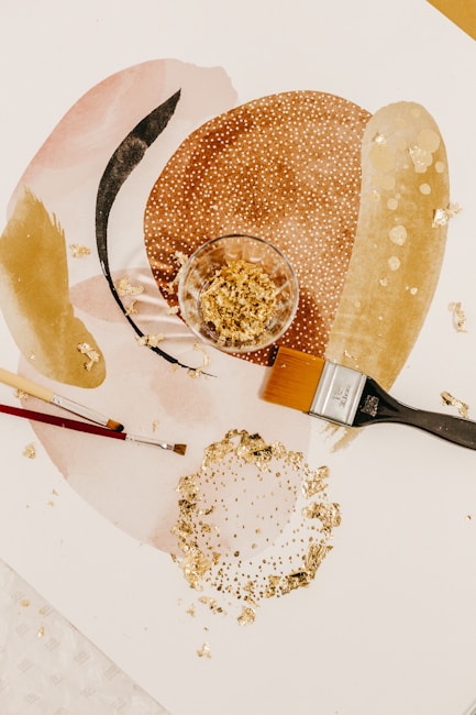 A collection of art supplies including two paintbrushes, a glass container with gold leaf fragments, and abstract art featuring soft colors and gold textures. The design includes overlapping ovals and circular patterns with shiny elements.