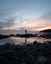 silhouette of person standing on rocky shore during sunset