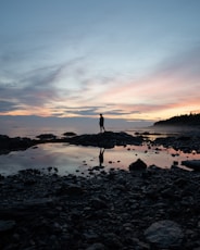silhouette of person standing on rocky shore during sunset