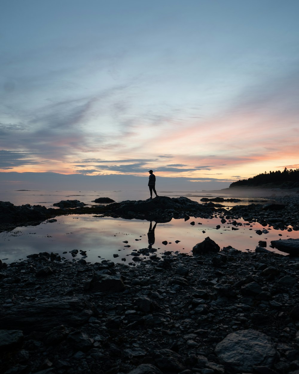 silhouette of person standing on rocky shore during sunset
