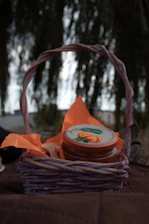 A small wicker basket contains a jar with a brightly colored label depicting apricots. The basket is lined with orange napkins. The background is blurred, with what appears to be a tree and some sky visible.