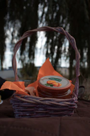 A small wicker basket contains a jar with a brightly colored label depicting apricots. The basket is lined with orange napkins. The background is blurred, with what appears to be a tree and some sky visible.