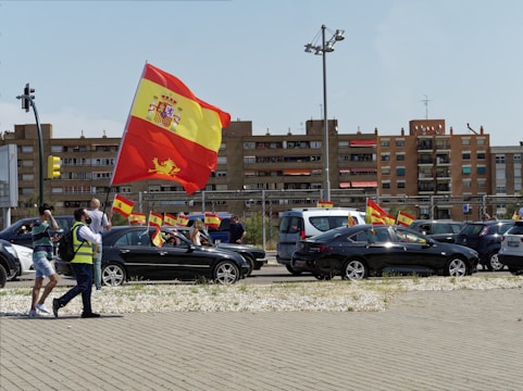 A street scene featuring a parade or protest with people and cars waving the Spanish flag. Several individuals are visible, including one in a reflective vest holding a large flag. Multiple vehicles display smaller flags. In the background, residential buildings and urban infrastructure are visible.