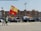 A street scene featuring a parade or protest with people and cars waving the Spanish flag. Several individuals are visible, including one in a reflective vest holding a large flag. Multiple vehicles display smaller flags. In the background, residential buildings and urban infrastructure are visible.