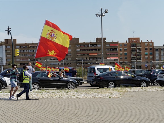 A street scene featuring a parade or protest with people and cars waving the Spanish flag. Several individuals are visible, including one in a reflective vest holding a large flag. Multiple vehicles display smaller flags. In the background, residential buildings and urban infrastructure are visible.