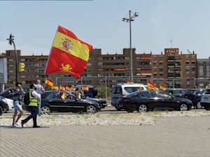A street scene featuring a parade or protest with people and cars waving the Spanish flag. Several individuals are visible, including one in a reflective vest holding a large flag. Multiple vehicles display smaller flags. In the background, residential buildings and urban infrastructure are visible.