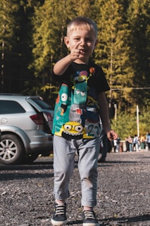 A young child stands pointing forward while wearing a colorful T-shirt featuring cartoon designs and light denim pants. The background shows a wooded area and a parked car, with soft sunlight illuminating the scene.