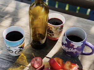 A wooden table is set with a brown glass bottle and three mugs filled with a dark red liquid. The mugs have different designs, including a bicycle theme, a festive theme, and a brand logo. Sliced red fruit, possibly plums or apples, is arranged on a small wooden cutting board in the foreground.