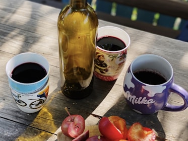 A wooden table is set with a brown glass bottle and three mugs filled with a dark red liquid. The mugs have different designs, including a bicycle theme, a festive theme, and a brand logo. Sliced red fruit, possibly plums or apples, is arranged on a small wooden cutting board in the foreground.