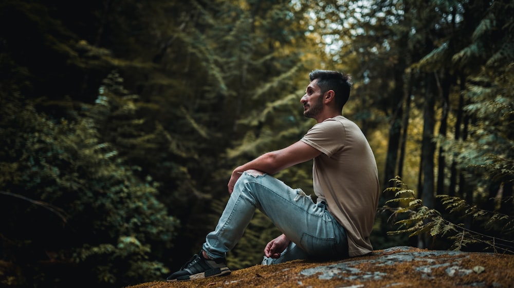 man in brown t-shirt and blue denim jeans sitting on brown wooden log during daytime