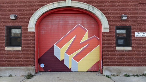 A red brick building features a large garage door with a bold, geometric mural of the letter 'M.' The mural uses a three-dimensional design with red and yellow colors and casts a shadow. There are two windows with black grills on either side of the door, and a 'Drivers Entrance' sign is visible.