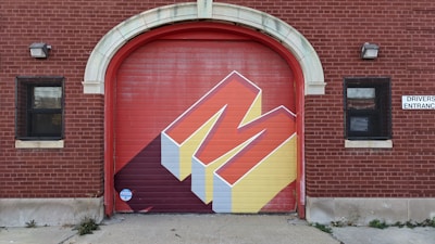 A red brick building features a large garage door with a bold, geometric mural of the letter 'M.' The mural uses a three-dimensional design with red and yellow colors and casts a shadow. There are two windows with black grills on either side of the door, and a 'Drivers Entrance' sign is visible.