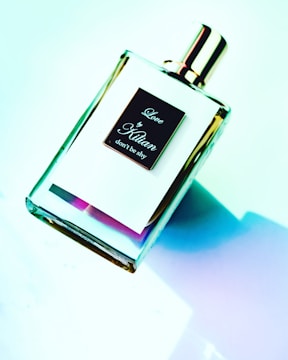 A perfume bottle labeled 'Love by Kilian, don't be shy' is shown against a gradient background with a mix of blue, green, and pink hues. The bottle has a reflective gold cap and a transparent design that reveals the liquid and a hint of color inside.