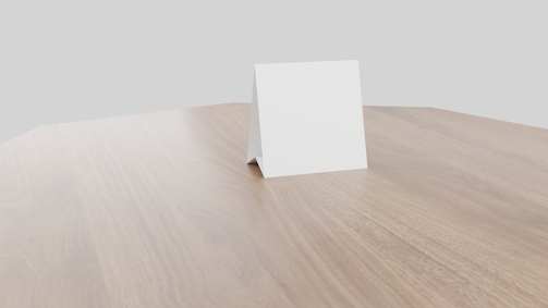 A blank white card is standing in a tent-like fashion on a smooth wooden surface. The tabletop has a light brown, natural wood grain texture, and the background is a plain, light grey color.