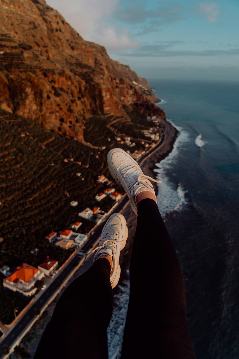 person in black pants and white sneakers sitting on brown rock formation near body of water