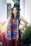A person wearing a graduation cap and vibrant stole stands confidently amidst colorful flowers and greenery. The stole reads 'Excellence in Diversity' and is decorated with bright, multicolored stripes. The person is dressed in a striped, sleeveless outfit. The background includes blurred elements, likely part of a garden or park setting with stone columns.